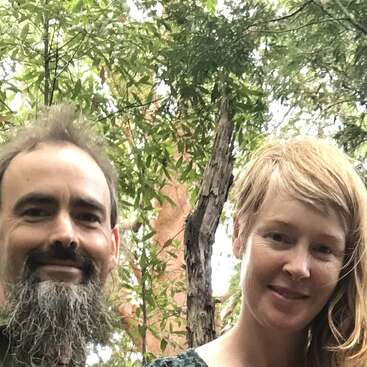 A man with a bushy beard and a smiling woman with light hair stand closely together outdoors, surrounded by lush green trees and natural forest scenery.