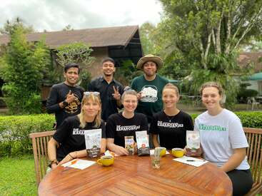 Siete personas posan alegremente al aire libre ante una mesa redonda de madera. Cuatro mujeres están sentadas con café y paquetes de productos. Tres hombres están de pie detrás, sonriendo con vegetación alrededor.