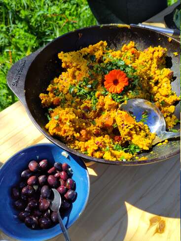In einer schwarzen Pfanne liegt ein leuchtend gelbes Gericht, garniert mit Kräutern und einer Orangenblüte. Daneben steht eine blaue Schüssel mit dunklen Oliven und einem Löffel.