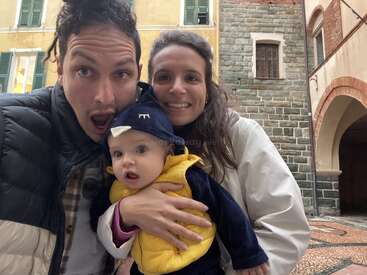 A joyful couple poses closely with their baby dressed in a cute animal costume. They smile, standing in a charming old European street with historic buildings.