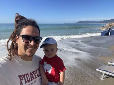 A smiling man with sunglasses and a child in a red shirt pose for a photo on a sunny beach, waves and mountains in the background.