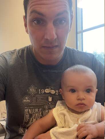 A man with short dark hair and a serious expression holds a baby with big eyes and light clothing, sitting indoors near a sunlit window.
