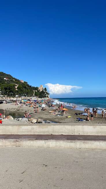 A lively beach scene with many people sunbathing and swimming. Blue sky, turquoise sea, green hills, and a castle-like building in the background. Summer atmosphere.
