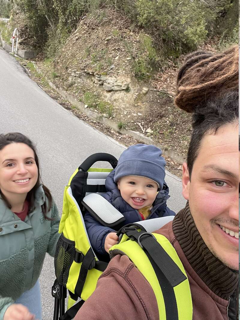 A happy family enjoys an outdoor walk. The father carries a smiling baby in a hiking carrier, mother walks beside them. Everyone looks cheerful and the surroundings are natural.