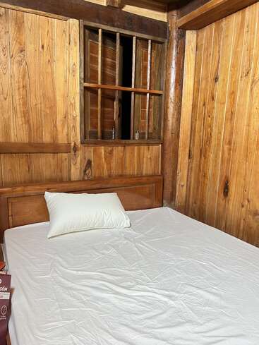 Um quarto simples de madeira apresenta uma cama de solteiro com lençol e travesseiro brancos. Há uma pequena janela com barras de madeira acima da cama, um ambiente rústico.