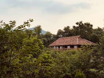 Uma casa de madeira rústica com telhado inclinado fica aninhada entre árvores verdejantes, cercada por uma floresta densa, uma atmosfera serena e montanhas ao fundo.