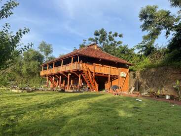 Uma tradicional casa de palafitas de madeira fica em um quintal verde exuberante, cercado por árvores e plantas, com uma pessoa trabalhando no jardim sob um céu azul.