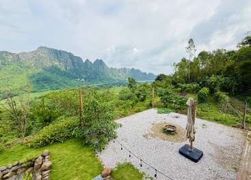 Um jardim tranquilo com pátio de cascalho e guarda-sol fechado tem vista para árvores verdes exuberantes, montanhas distantes e um céu nublado, criando uma paisagem tranquila e cênica.
