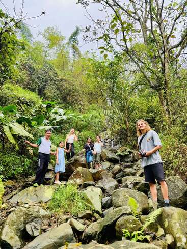A group of people stand on rocks in a lush, green forest. They appear to be hiking, exploring nature together, surrounded by trees and vegetation.