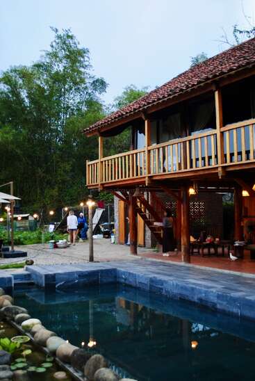This image shows a rustic wooden house with a red-tiled roof, an inviting swimming pool, people relaxing outside, surrounded by trees and cozy outdoor lights.