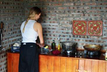 A woman with a braid washes dishes in a rustic kitchen with a brick wall, wooden counter, and pots on a stove, creating a cozy atmosphere.