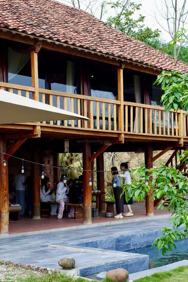 A wooden house with a red tiled roof, surrounded by greenery. People gather under the porch near a swimming pool, enjoying a relaxed outdoor atmosphere.