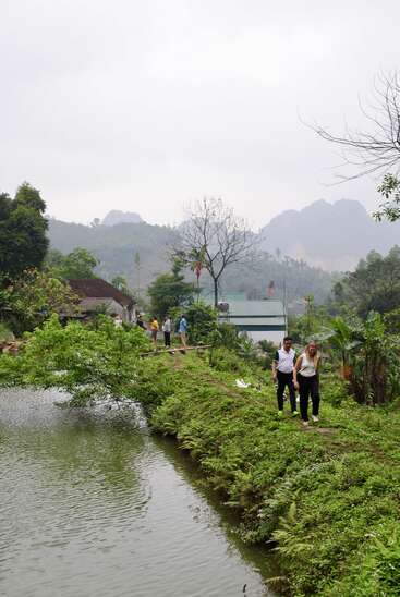 A group of people walk along a lush, green path beside a calm pond, surrounded by trees, houses, and distant misty mountains under a cloudy sky.