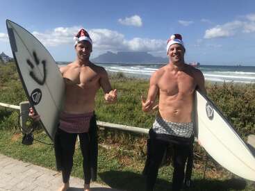 Zwei Männer mit Weihnachtsmützen stehen mit Surfbrettern in der Hand und ohne Hemd am Strand. Sie lächeln, machen Shaka-Zeichen und haben ihre Neoprenanzüge teilweise heruntergezogen, hinter ihnen sind Wellen zu sehen.