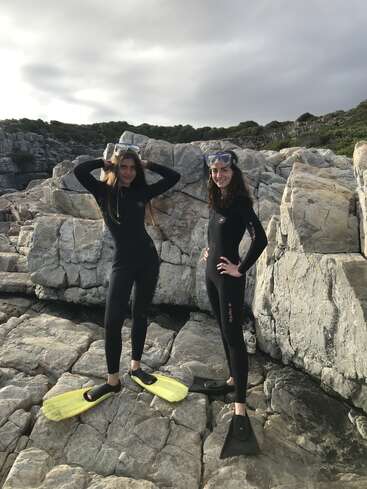 Zwei Frauen in Neoprenanzügen, mit Schnorchelmasken und Flossen stehen selbstbewusst auf felsigem Untergrund am Meer, lächeln und sind bereit für ein abenteuerliches Bad oder einen Tauchgang.
