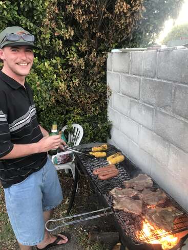 Ein lächelnder Mann grillt Steak, Würstchen und Mais auf einem Gartengrill. Er hält eine Zange und ein Getränk in der Hand und genießt einen entspannten Tag im Garten.