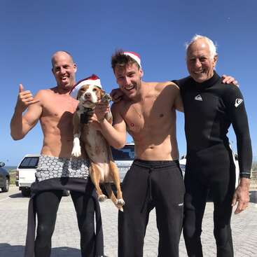 Três homens e um cachorro, todos usando chapéus de Papai Noel, posam alegres em uma praia ensolarada. Dois homens estão sem camisa, vestindo roupas de mergulho; um deles usa uma roupa de mergulho completa.