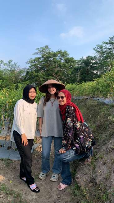 Trois femmes posent ensemble dans un jardin luxuriant, deux portant des hijabs, l'autre un chapeau traditionnel. Elles sourient, entourées de plantes vertes sous un ciel bleu.