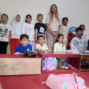 Un grupo de niños y una mujer adulta posan juntos en un aula. Algunos niños están sentados, otros de pie. Se ven mochilas en el suelo y en las estanterías.