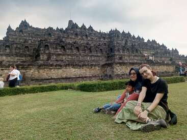 Três pessoas sentadas na grama verde, sorrindo juntas em frente ao antigo e majestoso Templo Borobudur, com outros turistas explorando o local histórico ao fundo.