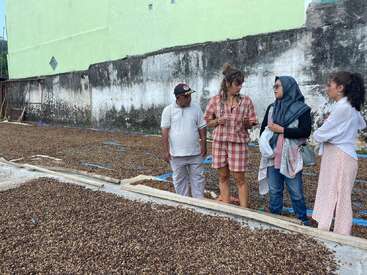 Quatro pessoas conversam ao lado de grandes camas de secagem cobertas de grãos de café, colocadas ao ar livre ao lado de uma parede de concreto desgastada pelo tempo e de um prédio verde-claro.