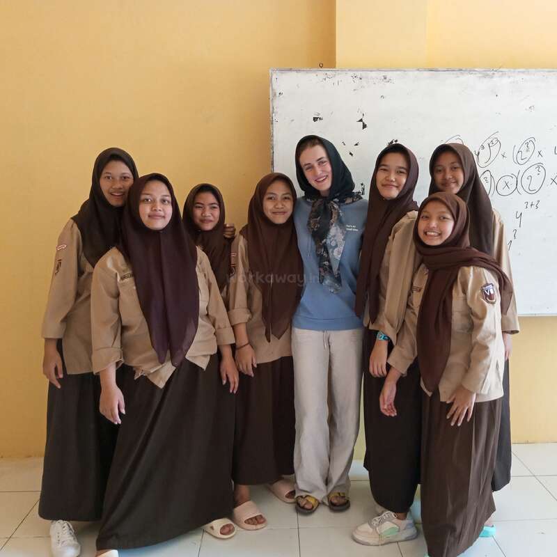 Um grupo de sete meninas com uniformes marrons e lenços de cabeça está sorrindo com uma mulher em uma sala de aula, em frente a um quadro branco com equações matemáticas.