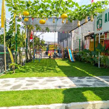A small, vibrant playground with green grass, slides, and climbing structures. Overhead, vines with yellow gourds provide shade. Colorful bunting and signs decorate the area.