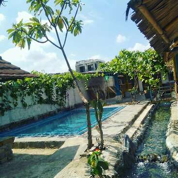A serene outdoor courtyard featuring a refreshing swimming pool, lush green vines, a flowing stream, tropical trees, rustic huts, and clear blue skies on a sunny day.