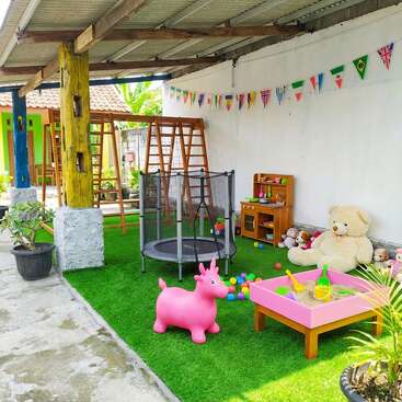 A colorful outdoor play area features artificial grass, a trampoline, swings, a sandbox, plush toys, play kitchen, bunting flags, and various vibrant children's toys and decorations.