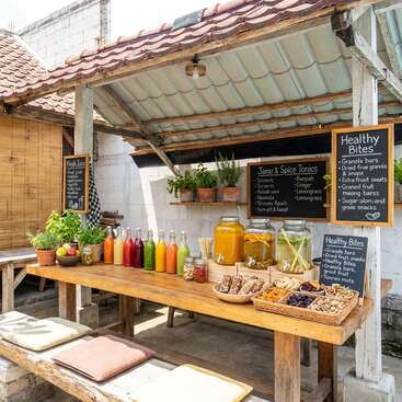 A rustic juice and snack stall offers colorful fresh juices, herbal tonics, granola bars, dried fruit, and healthy snacks, with handwritten chalkboard menus and potted herbs.