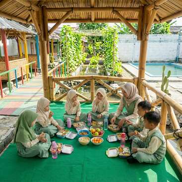 A group of children and a teacher in uniforms sit together outdoors under a bamboo shelter, sharing a healthy meal picnic on a green mat, smiling.