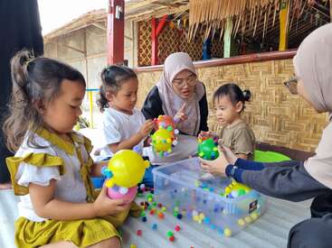 Três crianças e dois adultos estão sentados no chão, brincando com brinquedos coloridos e bolas. Os adultos envolvem as crianças numa atividade divertida e educativa.