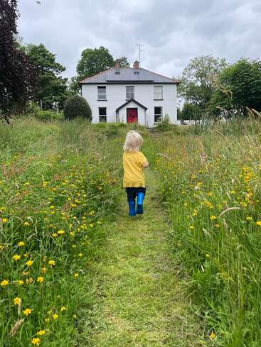 Un petit enfant chaussé de bottes bleues et vêtu d'une chemise jaune marche le long d'un chemin herbeux bordé de fleurs sauvages, en direction d'une maison blanche dotée d'une porte rouge vif.