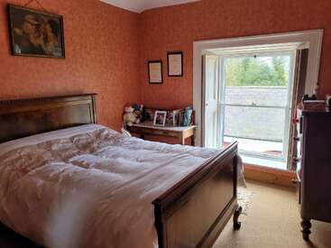 This cozy bedroom features a wooden bed, orange wallpaper, a dresser, framed pictures, open window, bedside table with books, certificates, and plush toys.
