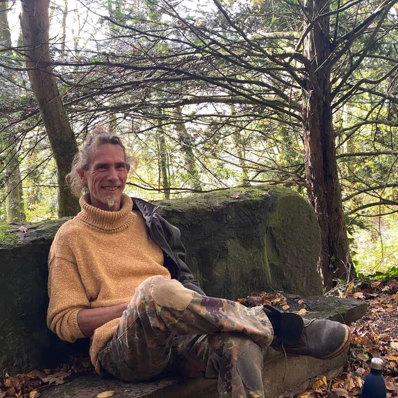 The image depicts a man sitting on a moss-covered stone bench in a wooded area, surrounded by trees and fallen leaves, with a serene and natural atmosphere.