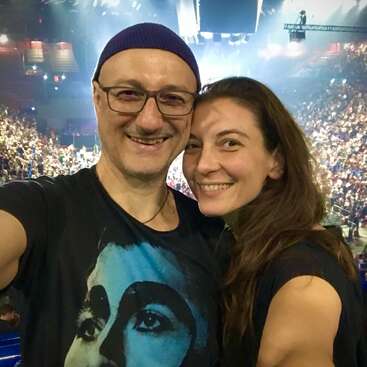 A happy couple takes a selfie at a crowded concert. They are smiling, enjoying the lively atmosphere, surrounded by an excited audience under bright lights.