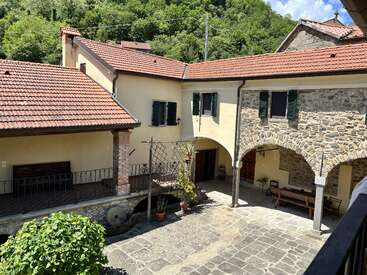 This charming courtyard features stone arches, yellow walls, terracotta roof tiles, green shutters, outdoor seating, potted plants, and is surrounded by lush hillside greenery. Peaceful ambiance.