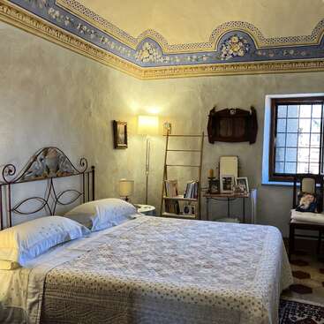 This cozy bedroom features a wrought iron bed, vintage decor, a patterned ceiling border, warm lighting, a bookshelf, and a window letting in natural light.