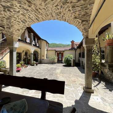 A charming stone courtyard with arches, potted plants, and staircases. Sunlight highlights rustic architecture. A wooden bench and table sit shaded in the foreground. Peaceful atmosphere.