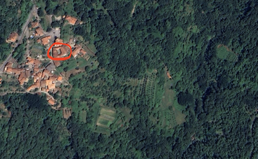 Dieses Luftbild zeigt ein ländliches Dorf inmitten eines dichten grünen Waldes. Ein roter Kreis hebt ein bestimmtes Gebäude innerhalb der Häusergruppe hervor, um seine Lage zu betonen.