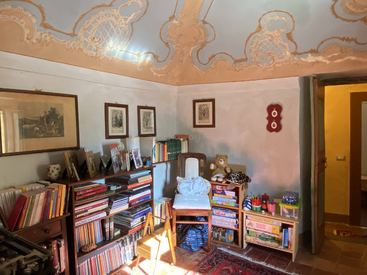 In diesem gemütlichen Zimmer gibt es verschnörkelte Decken, Bücherregale voller Bücher, gerahmte Kunst, Kinderspielzeug, Brettspiele, einen Holzstuhl und sanftes Sonnenlicht, das alles warm beleuchtet.