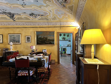 Ein warm beleuchtetes Esszimmer im Vintage-Stil mit gelben Wänden, einer verzierten Decke, klassischen Gemälden, Holzmöbeln, Dekoartikeln und einer leuchtenden gelben Lampe, das mit einer Küche verbunden ist.