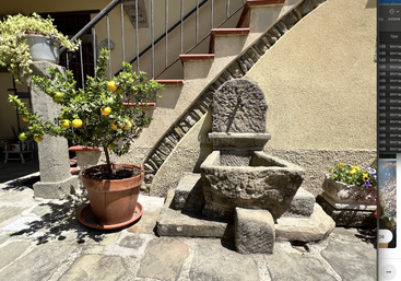 Ein getöpferter Zitronenbaum, ein alter Steinbrunnen und farbenfrohe Blumen stehen in einem sonnenbeschienenen Innenhof unter einer Treppe mit Terrakottastufen und Eisengeländer.