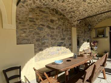 Dieses Bild zeigt eine gemütliche Steinterrasse mit einem Holztisch, Bänken und Stühlen. Es gibt dekorative Gegenstände und Sonnenlicht, das auf den Platz fällt.