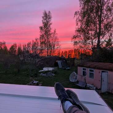 Una persona descansa en una azotea, con los pies visibles en primer plano, contemplando un patio trasero rústico al atardecer, con tonos rosados y morados que iluminan el cielo.
