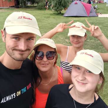 Cuatro personas sonrientes posan para hacerse un selfi al aire libre; todas llevan gorras beige de «ZS Rozítek». Al fondo se ve una tienda de campaña y césped verde, lo que sugiere que se trata de una acampada.