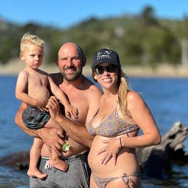 Une famille souriante de trois personnes se tient ensemble à la plage. L'homme tient un jeune garçon dans ses bras et la femme, enceinte, porte des lunettes de soleil et un chapeau. Journée ensoleillée.