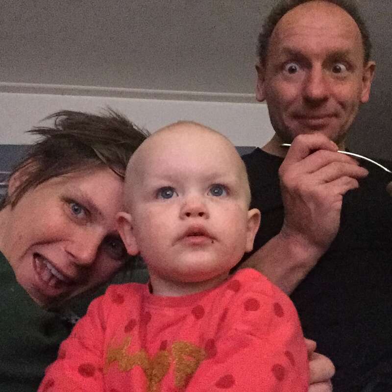 The image shows a family of three posing for a selfie, with a baby in the foreground and a man and woman behind them, all smiling.