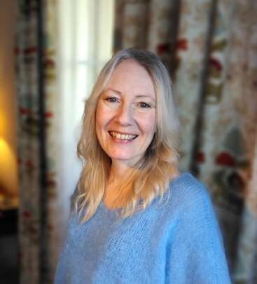 Eine lächelnde Frau mit langen blonden Haaren steht drinnen und trägt einen hellblauen Pullover. Sanftes Licht und gemusterte Vorhänge sorgen für eine warme, einladende und fröhliche Atmosphäre.