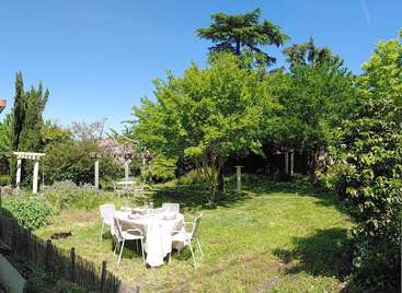 Ein sonniger Garten mit üppig grünen Bäumen, ein runder Tisch für vier Personen mit weißer Tischdecke, Stühle, eine Pergola und eine entspannte Atmosphäre.